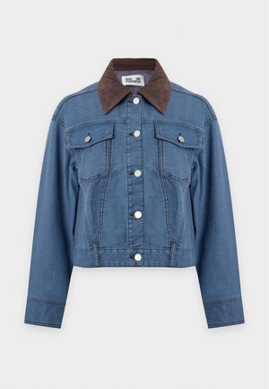 Femme debout portant une veste en jean bleue à col foncé, un pantalon large assorti, un haut crème boutonné et des chaussures à talons noirs.