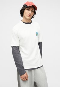 Νέος άνδρας που φοράει λευκό T-shirt με ένα μικρό μπλε λογότυπο καρτούν χελώνας, από πάνω από γκρι μακρυμάνικο πουκάμισο και κόκκινο καπέλο.