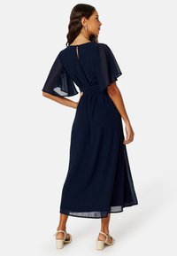 Námořnické modré maxi šaty s průhlednými volánovými rukávy, přiléhavým pasem a rozparkem. Disponují detailem s klíčovou dírkou na zadní stránce a splývavou tkaninou.