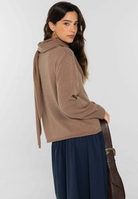 Jersey marrón de punto con capucha y detalle de lazo en la parte trasera, combinado con una falda azul marino plisada. Modelo sosteniendo un bolso de cuero marrón.