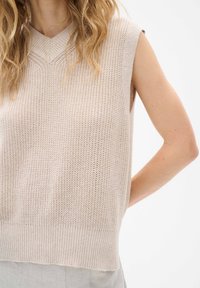 Beige stickad ärmlös topp med V-hals, utrustad med öppna stickmönster och ribbad nederkant. Diskret textur förbättrar designen.