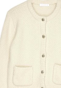 Kremo spalvos mezginis kardiganas su apvalia iškirpte, dviem priekiniais kišenėmis ir šešiais dekoratyviniais, spiralės formos metaliniais mygtukais. Tekstūruota medžiaga.