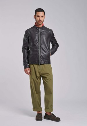 Man staat met één hand in zijn zak en draagt een zwarte leren jas, olijfgroene opgerolde broek en donkerbruine casual schoenen.