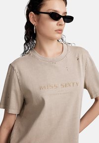 Camiseta de algodón beige con detalles desgastados y el logotipo bordado "MISS SIXTY". Modelo con gafas de sol negras y un corte holgado.