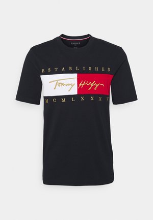 Černé bavlněné tričko s kulatým výstřihem, s bílým a červeným horizontálním pruhem. Zlatý text "ESTABLISHED", "Tommy Hilfiger" a římské číslice.