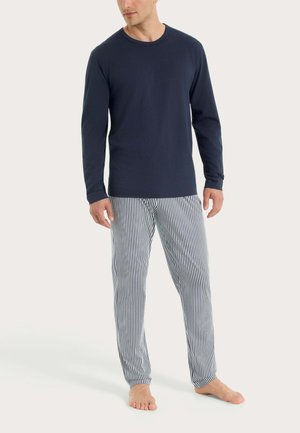 Uomo con una maglia a maniche lunghe blu navy e pantaloni a righe verticali bianchi e blu navy, in piedi a piedi nudi su uno sfondo plain.