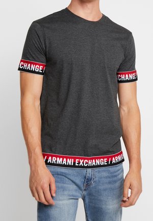 Mand iført mørkegrå kortærmet T-shirt med rød, sort og hvid Armani Exchange-brandet kant på ærmer og kant, kombineret med blå jeans.