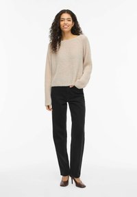 Pull en tricot beige clair avec manches larges, associé à un pantalon noir à jambes droites et des talons hauts marron. Texture lisse et coupe décontractée.