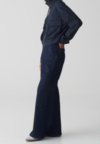 Mörkblå zip-up jacka med hög krage och två frontfickor; ihop med mörka denimbyxor med vida ben och grå skor.