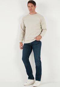 Sudadera de color beige claro con cuello redondo y mangas ranglán, combinada con vaqueros azul oscuro y zapatillas blancas, posando frente a un fondo liso.