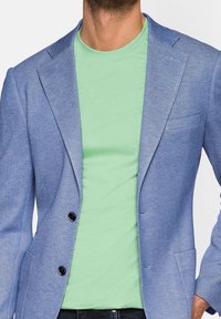 Světle modrý blazer s texturovaným povrchem a klopovými límci, nošený přes mintově zelené tričko. Obsahuje dva černé knoflíky a boční kapsy.