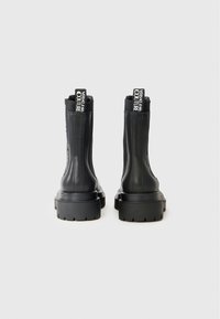 Black ankle boots with a smooth, glossy finish, chunky sole, and contrasting branded pull tabs at the back. Reinforced stitching along sides.