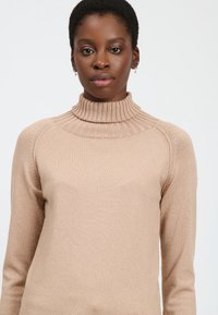 Maglione a collo alto beige a coste con maniche lunghe, caratterizzato da un design con collo alto e una texture liscia e leggermente lucida.