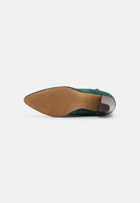 Chaussure en daim vert à bout pointu vue de la semelle, montrant une semelle extérieure lisse beige avec une texture antidérapante près de l’avant-pied et un talon carré.