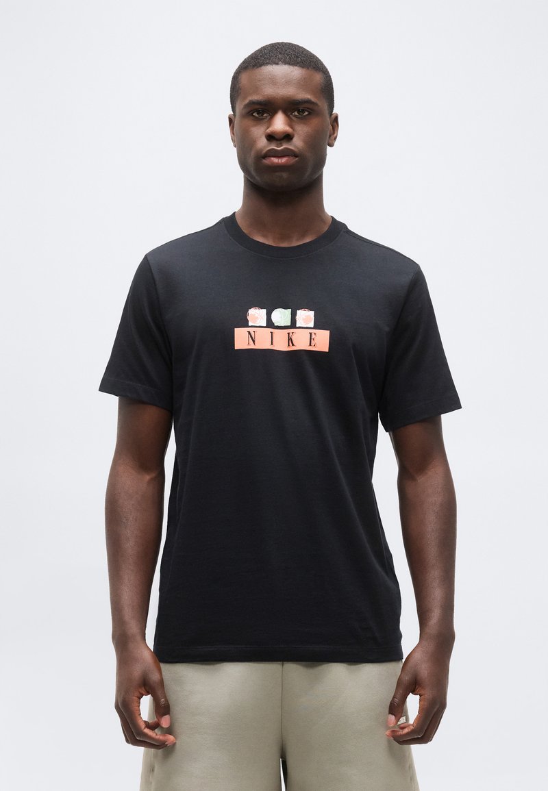 Άνδρας που φοράει μαύρο μπλουζάκι Nike με ροδακινί και πράσινα αφηρημένα σχέδια πάνω από το λογότυπο, συνδυασμένο με ανοιχτό μπεζ σορτς.