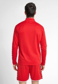Rode lange mouw atletische top van een gestructureerde stof, met hoge kraag en zwarte zijaccenten. Draagt een bijpassende rode short.