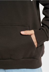 Mørkebrun sweatshirt med kangaroo-lomme, ribbede manchetter og kant. Glat tekstur med synlige syningsdetaljer. Hånd delvist i lommen.