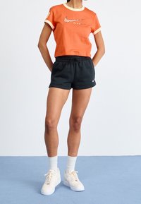 Oranje kort T-shirt met crèmekleurige bies en Nike-logo; zwarte elastische korte broek; crèmekleurige sneakers; witte sokken. Eenvoudig sportief ontwerp.