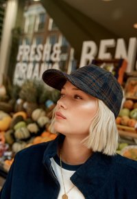 Blauer kariertes Baseball-Cap mit gebogenem Schirm und strukturierter Stoffoberfläche. Dunkle Jacke über einem weißen Hemd, vor einem Hintergrund aus Obst und Gemüse.