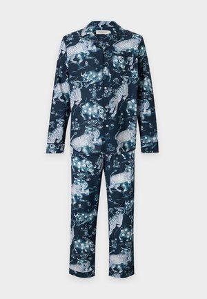 Homme debout pieds nus portant un pyjama bleu foncé avec un motif de tigre blanc et floral sur un fond clair uni.