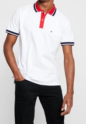 Homme portant un polo blanc avec col rouge et bordure marine, pantalon noir, une main dans la poche, debout devant un fond uni.