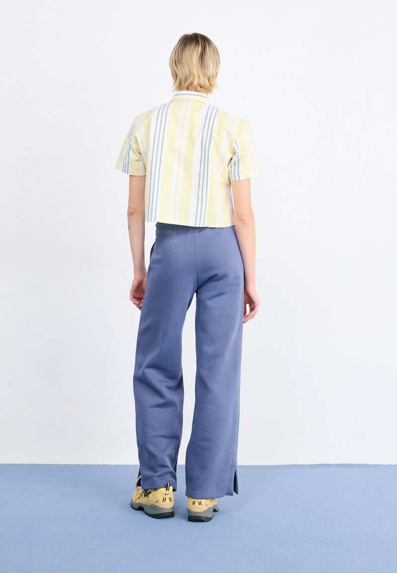 Gekürztes, gelb-grün gestreiftes Hemd, kombiniert mit locker sitzenden blauen Hosen und gelben Turnschuhen mit schwarzen Akzenten. Einfaches, lässiges Design.