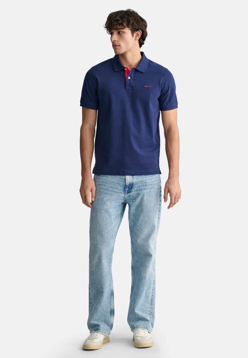 Junger Mann schaut nach links, trägt ein navyblaues Poloshirt mit roten Knöpfen, hellblaue Jeans und weiße Sneakers vor einem hellgrauen Hintergrund.