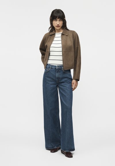 Γυναίκα με καφέ μπουφάν, λευκό πουκάμισο με ρίγες, φαρδιά μπλε τζιν και σκούρα καφέ παπούτσια, που στέκεται μπροστά σε απλό φωτεινό φόντο.