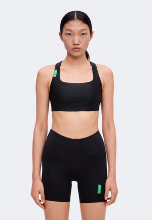 Jeune femme debout face à l’avant portant un soutien-gorge de sport noir et un short de cyclisme noir taille haute avec des étiquettes vertes sur chaque vêtement.
