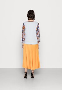 Top azzurro chiaro con maniche a sbuffo decorate; abbinato a una gonna arancione fluente e plissettata. Scarpe nere con tacchi alti su un pavimento grigio.