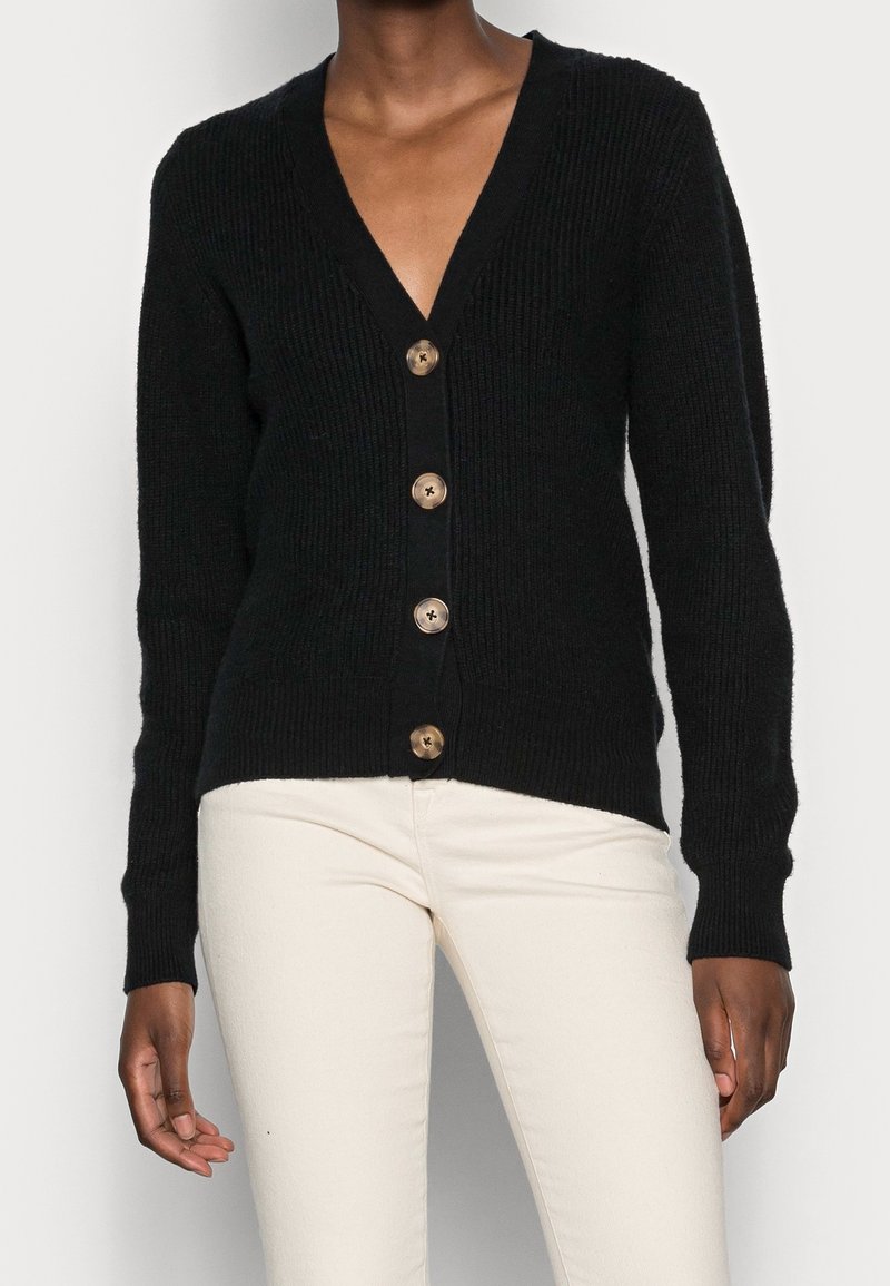Cardigan noir en maille côtelée avec quatre gros boutons marron, porté avec un pantalon ajusté écru.