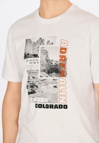 Vit bomull t-shirt med en grafisk design som har svart och orange text, flera fotocoller och geografiska koordinater.