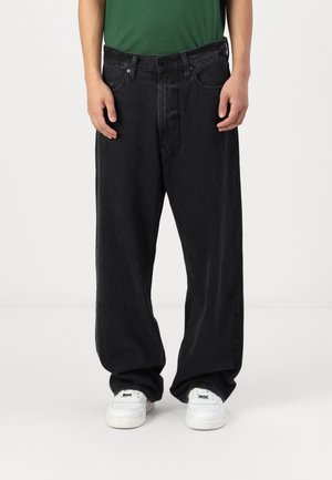 Black wide-leg jeans with a textured finish, featuring front pockets, belt loops, and a classic five-pocket design. White sneakers visible.