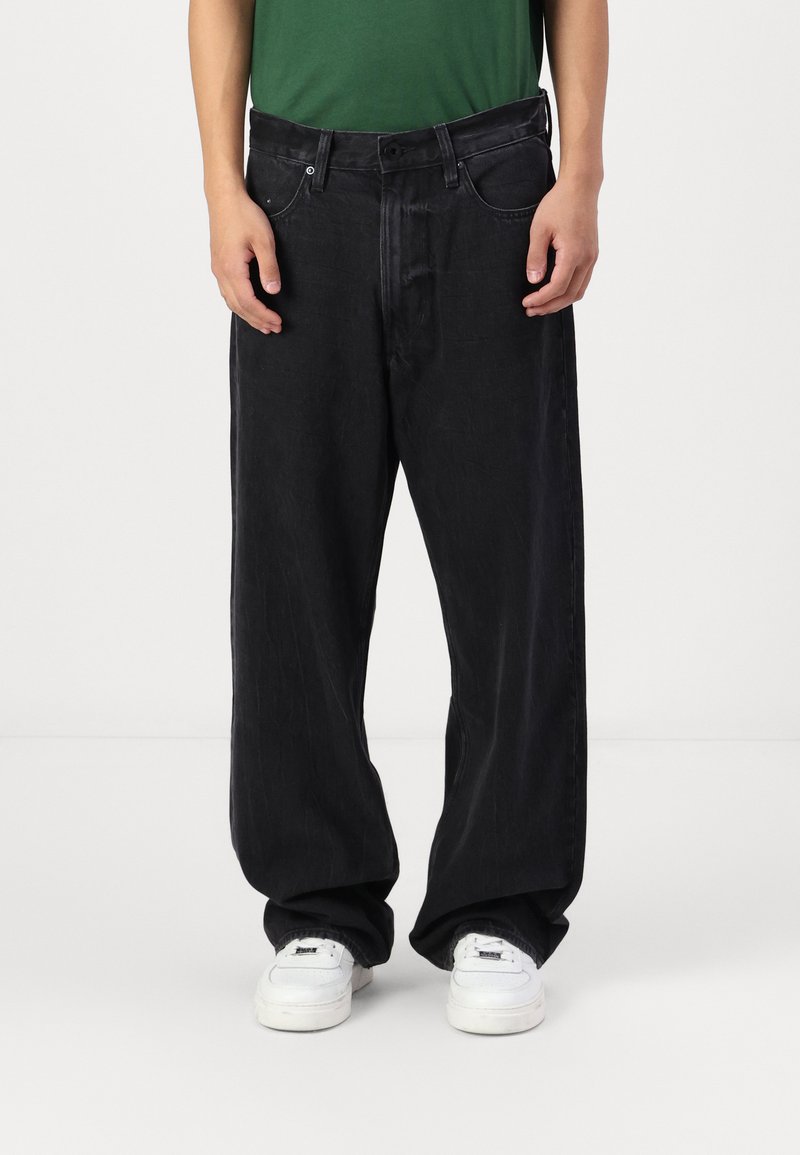 Jeans negre cu croială largă și finisaj texturat, dotate cu buzunare frontale, bucle pentru curea și un design clasic cu cinci buzunare. Adidași albi vizibili.