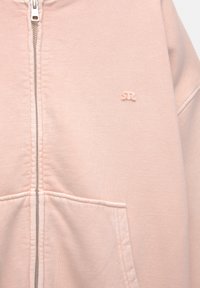 Lichtroze zip-up hoodie met een voorzak en een klein geborduurd logo op de borst, met zichtbare stoftextuur en ritsdetail.
