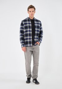 Camisa de franela de cuadros en azul marino y beige con cierre de botón, combinada con jeans grises y zapatillas deportivas negras. Textura de tela suave.