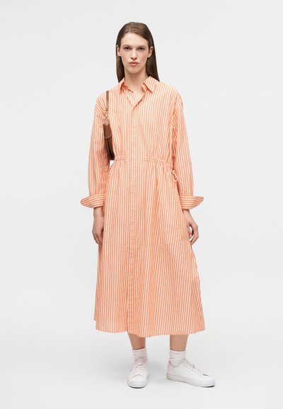 Πορτοκαλί και λευκό φόρεμα-πουκαμίσα με μακριές ρίγες, γιακά με κουμπιά, ζωνάκι στη μέση και μακριά μανίκια, συνδυασμένο με λευκά παπούτσια.