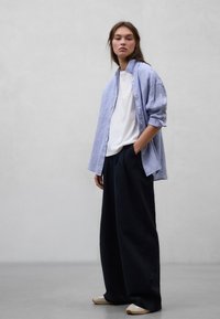 Ung kvinde iført løs lyseblå skjorte over en hvid t-shirt og vide sorte bukser, stående mod en ensartet lysegrå baggrund.