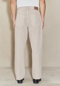 Pantalon en denim beige à coupe droite, avec deux poches arrière et un patch texturé sur la ceinture, accompagné de chaussures noires.