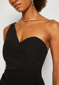 Femme portant une robe noire asymétrique avec un collier chaîne serpent doré et des boucles d'oreilles pendantes dorées sur fond uni.