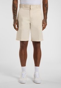 Beige Baumwollshorts mit flachem Frontschnitt, Seitentaschen und knielanger Passform, kombiniert mit weißen Sneakers und knöchellangen Socken.