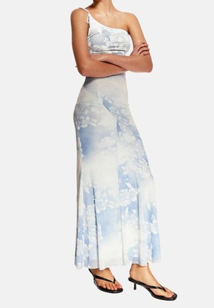 Femme portant une robe maxi à imprimé floral bleu et blanc à une épaule, avec des sandales noires à talons bas, debout les bras croisés.
