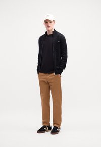 Ung mand iført hvid kasket, sort lynlås kabelstriktrøje, sort skjorte, beige bukser og sorte sneakers med hvide striber, som står med hænderne i lommen.