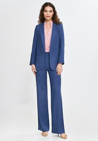 Ciemnoniebieski garnitur z dopasowaną marynarką i szerokimi spodniami, w zestawieniu z jasnoróżową bluzką z kołnierzykiem. Gładki materiał i dopasowany krój.