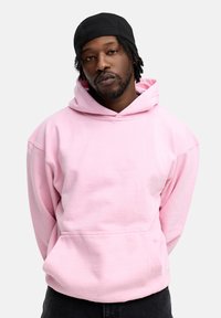 Pink hooded sweatshirt made of soft fabric with a front pocket. Features a drawstring hood and relaxed fit, worn with dark jeans.