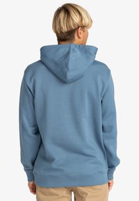 Hombre con un sudadero azul con capucha y pantalones beige, de espaldas a la cámara sobre un fondo blanco liso.