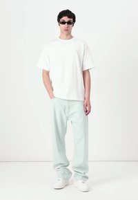 Férfi fekete napszemüvegben, fehér, oversized pólóban, világoskék bőrnadrágban és fehér sportcipőben, egy sima fehér háttér előtt áll.