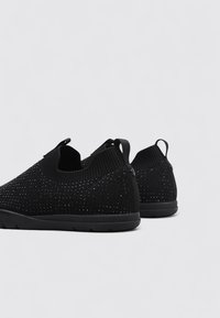 Zapatos negros sin cordones hechos de una tela de malla transpirable con una superficie texturizada, que cuentan con una suela flexible y una pestaña en el talón.