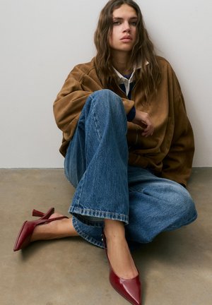 Hosszú hajú fiatal nő ül a padlón, túlméretezett barna dzsekit, bő kék farmert és piros, magas sarkú, szíjas cipőt visel.