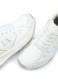 White athletic sneakers with textured leather, subtle gray accents, rounded toe, and flat laces. Logo 'LA' printed on the side.
